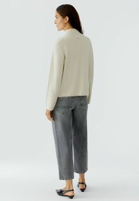 Beige Strickpullover mit hohem Kragen, kombiniert mit hochgeschnittenen grauen Jeans mit Gesäßtaschen und lockerer Passform. Schwarze Slipper.