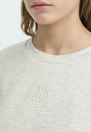 Close-up af en person iført en lysegrå skjorte med et diskret præget logo på brystet.