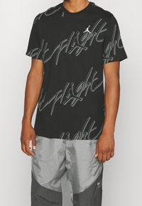 Homme portant un T-shirt graphique noir avec du texte "Flight" répété et des logos Jumpman, associé à un pantalon Nike argent et noir.