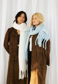 Twee vrouwen dragen lange, bruine suède jassen. De ene heeft een witte sjaal, de andere een lichtblauwe sjaal met rafelige uiteinden en een label met de tekst "Arctic Fox."