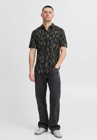 Chemise noire à manches courtes avec un motif de feuilles blanches, col boutonné, associée à un jean foncé et des baskets claires. Design minimaliste.