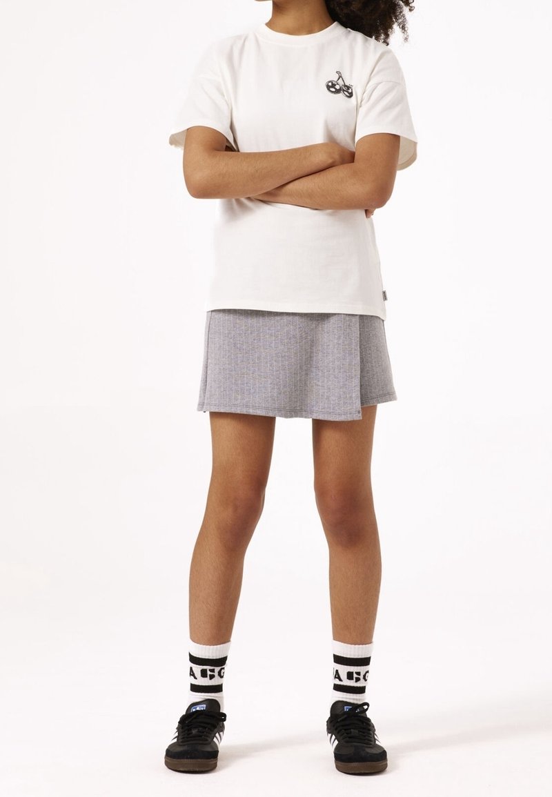 Enfant aux cheveux bouclés, debout les bras croisés, portant un t-shirt blanc avec un motif de vélo, une jupe grise, des chaussettes blanches et des baskets noires.