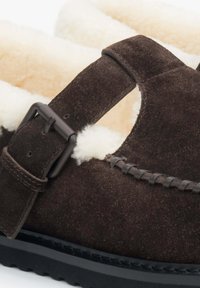 Hnědé semišové boty s beránkovou podšívkou, nastavitelným páskem a černou gumovou podrážkou. Obsahují prošité detaily podél okrajů.