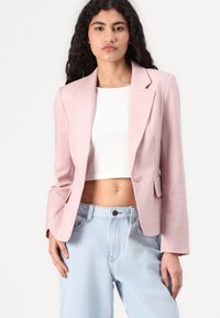 Světle růžový blazer s klopovým límcem, jednobotonovým zapínáním, předními kapsami a dlouhými rukávy. Nosený přes bílý crop top a světle modré džíny.