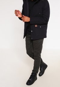 Svart vadderad jacka med huva, med dragkedja framtill och tryckknappar. Matchad med mörkgröna cargobyxor och svarta högklackade sneakers.