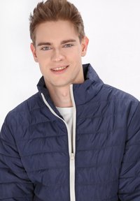 Marineblaues Steppjacke mit gestepptem Design, hohem Kragen und einem weißen Reißverschluss. Getragen über einem weißen Hemd, das eine glatte, glänzende Stofftextur aufweist.