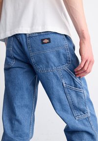 Pantalones cargo de denim azul claro con múltiples bolsillos, costuras visibles y una etiqueta negra en el bolsillo trasero. Ajuste regular y diseño de tiro medio.