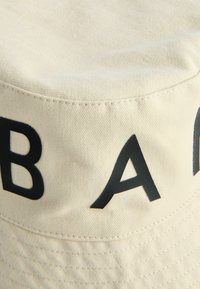 Beige Buckethat aus Stoff mit sichtbaren Nähten. Das Band hat große schwarze Buchstaben. Glatte Textur und runde Form.