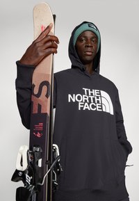 Zwarte hoodie met witte "The North Face" logo, gecombineerd met ski's met een bruin en zwart ontwerp. Een teal gebreide beanie compleet de look.