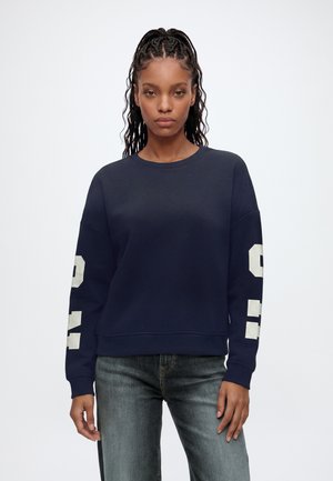 Frau mit geflochtenem Haar trägt ein marineblaues Sweatshirt mit weißen Blockzahlen an den Ärmeln und ausgewaschene blaue Jeans und steht vor einem schlichten weißen Hintergrund.