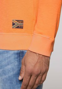 Oranje sweatshirt met ribgebreide manchetten en een gestructureerde afwerking. Bevat een blauw en geel Camp David logo-patch op de voorkant.