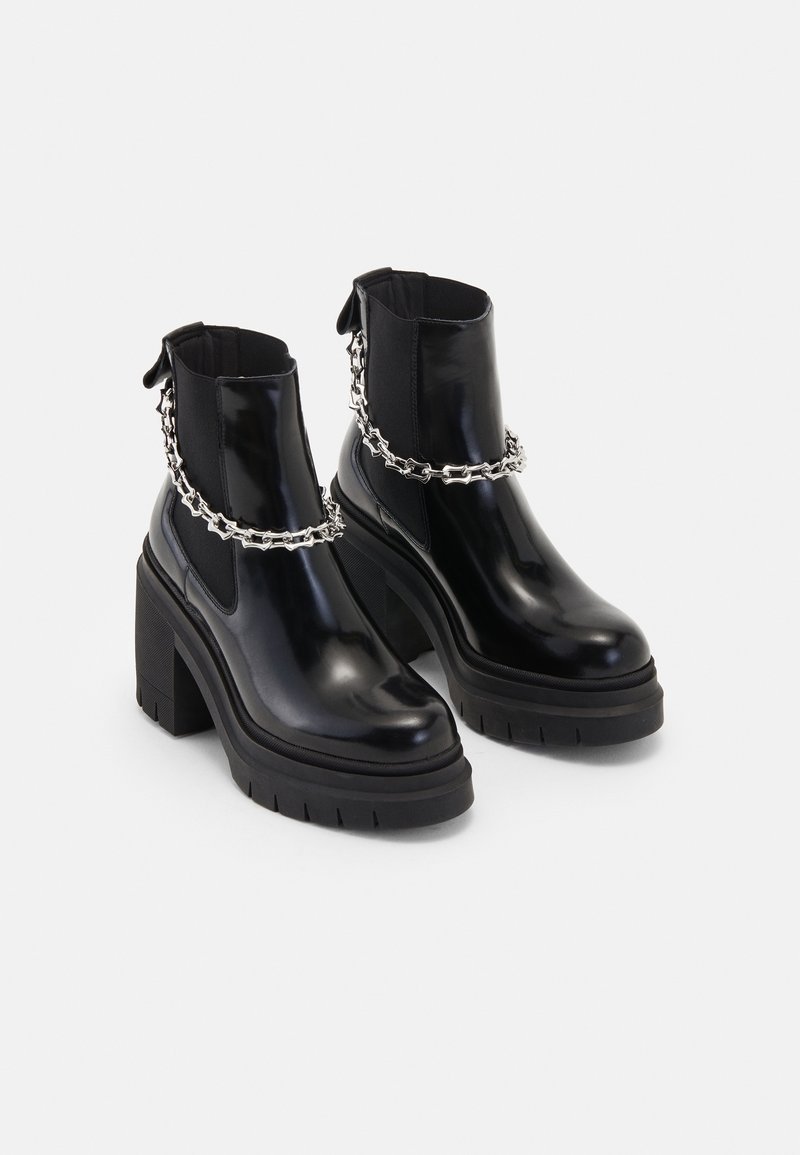 Sorte ankelstøvler med chunky hæl og en blank finish, elastiske sidespaneler og en sølvfarvet kæde rundt om anklen. Tykk sål med riller.