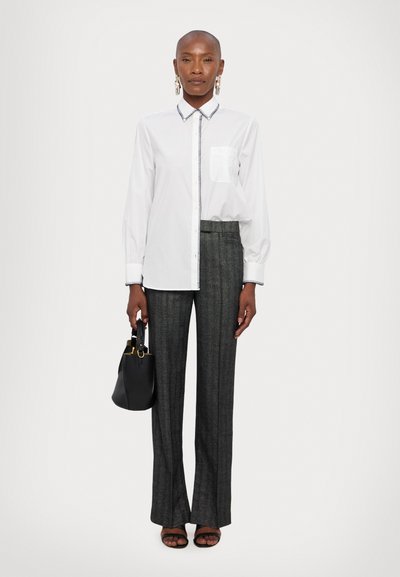 White button-up shirt with a pocket, paired with dark gray, pinstriped, tailored pants. Model holds a small black handbag.