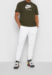 Hombre vistiendo una camiseta Nike Air verde oliva, pantalones jogger blancos y zapatillas multicolores, de pie con las manos en los bolsillos.