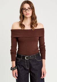 Brun, ribbet off-shoulder top med lange ærmer, parret med mørke denimbukser og et sort bælte. Bemærkelsesværdige accessories inkluderer et sort armbånd.