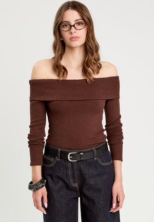 Top marrón de canalé con off-shoulder y mangas largas, combinado con pantalones de mezclilla oscuros y un cinturón negro. Los accesorios destacados incluyen una pulsera negra.