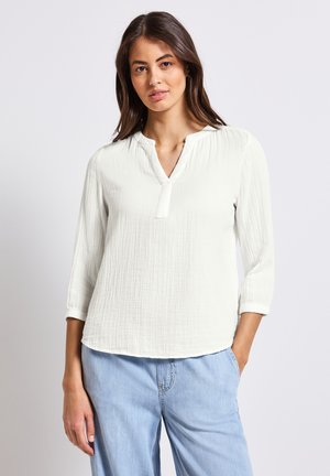 Blouse blanche texturée avec un décolleté en V, des manches trois-quarts et une coupe décontractée, associée à un pantalon bleu clair.