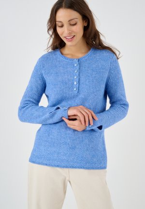 Donna che indossa un maglione azzurro chiaro di maglia con bottoni e maniche lunghe e pantaloni beige, guarda verso il basso e sorride dolcemente.
