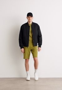 Crna bomber jakna, maslinasto zelena majica, usklađene kratke hlače, bijele tenisice i crna kapa. Glatka tkanina, jednostavan dizajn, minimalistički stil.