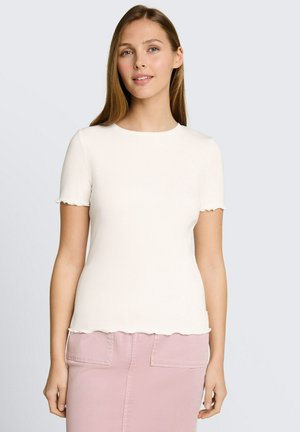 Młoda kobieta ubrana w biały, żeberkowany T-shirt z krótkim rękawem i jasnoróżową spódnicę, stojąca na jednolitym jasnoszarym tle.