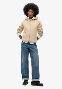 Felpa beige con zip, con cordini e tasche frontali, indossata sopra una camicia bianca con colletto; abbinata a jeans oversize azzurri e scarpe nere.