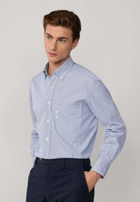 Hombre vestido con una camisa de rayas azul y blanca de manga larga con botones y pantalones oscuros, con una mano en el bolsillo, mirando de lado.