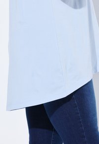 Top tunica azzurro chiaro su jeans attillati blu scuro su una persona, mostrata dalla vita in giù su uno sfondo pallido.