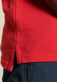 Polo shirt rouge en tissu texturé avec des fentes sur les côtés à l'ourlet, associé à un pantalon bleu foncé à motif à carreaux.