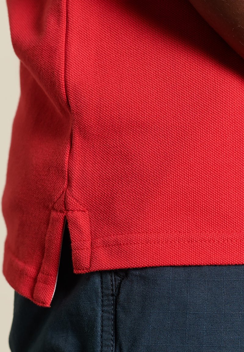 Polo shirt rouge en tissu texturé avec des fentes sur les côtés à l'ourlet, associé à un pantalon bleu foncé à motif à carreaux.