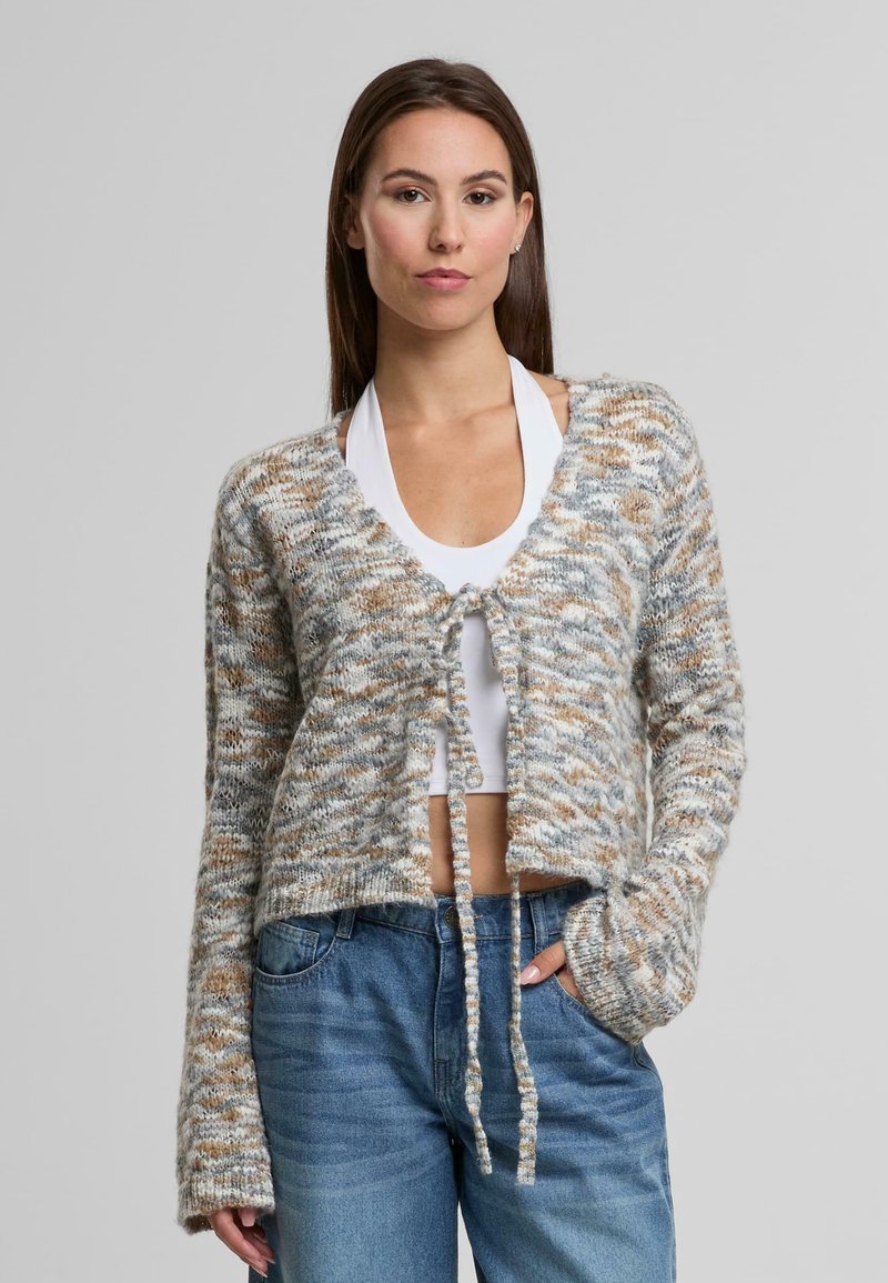 Háčkovaná kardigan v šedých, bílých a hnědých odstínech. Má zkrácený střih, otevřený přední díl se zavazovacími prvky a dlouhé rukávy.