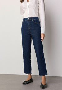 Tamsiai mėlynos aukšto liemens džinsai tiesaus kirpimo, su priekyje esančiu sagos užsegimu ir diržo kilpelėmis, derinami su balta marškinių palaidine.