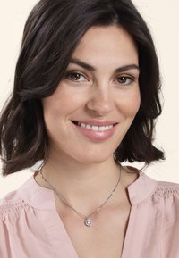 Collana d'argento con un pendente rotondo e una pietra trasparente, appoggiata su un décolleté con una blusa rosa chiaro e onde morbide nei capelli.