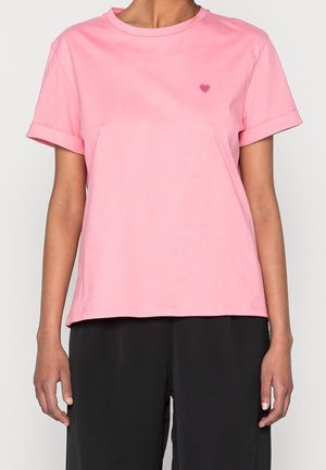 Kobieta nosząca jednolitą różową koszulkę z krótkim rękawem z podwiniętymi mankietami i małym haftowanym sercem na lewej piersi, w połączeniu z czarnymi spodniami.