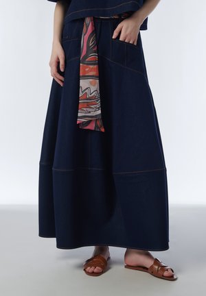 Vrouw draagt een lange donkerblauwe jeansrok met grote opgestikte zakken, een stoffen riem met patroon en bruine open-toe platte sandalen, staand op een eenvoudige vloer.