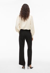 Kvinne med langt krøllete hår iført en kremfarget bluse, svarte skreddersydde bukser og svarte høye hæler, vendt bort fra kameraet mot en hvit bakgrunn.