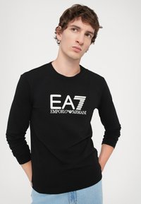 Fekete, hosszú ujjú póló, kerek nyakkivágással, amelyen hímzett, fehér "EA7 EMPORIO ARMANI" logó található az elején. Puha pamut textúra.