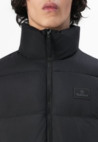 Sort dunjakke med høj krave, der har lynlås foran og et Timberland-logo senet på brystet. Glat, mat overflade.