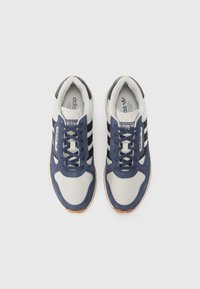 Adidași cu un exterior din suede albastru și plasă gri, cu dungi negre și o talpă din cauciuc de tip gumă. Șireturile și limba sunt în nuanță de albastru potrivit.