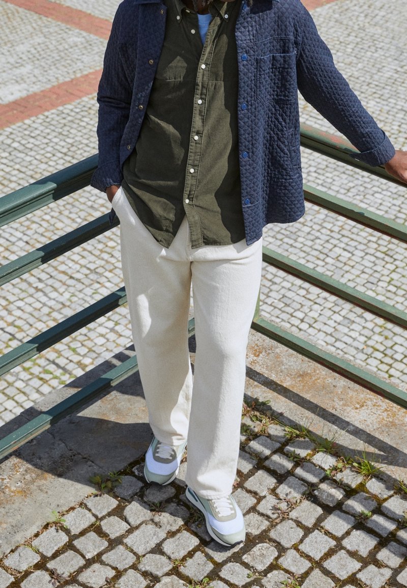 Personne appuyée sur une rambarde métallique verte en plein air, portant un pantalon blanc, des baskets blanches et grises, une chemise olive et une veste matelassée bleue.