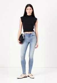 Haut noir sans manches, jean skinny bleu clair et ballerines crème. Le mannequin tient une pochette noire. Design simple et ajusté.