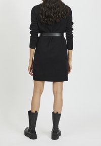 Abito nero lavorato a maglia con vita a cinturino, polsini e orlo a costine, abbinato a stivali neri in pelle al ginocchio. Design semplice e aderente.