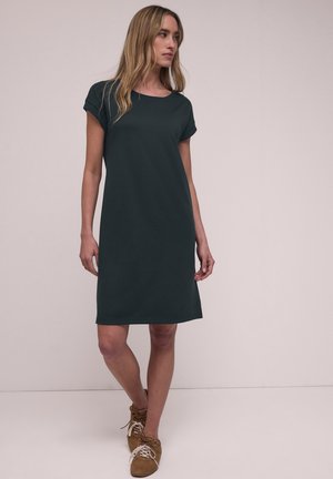 Femme aux longs cheveux blonds portant une robe courte verte foncée à manches courtes jusqu'aux genoux et des chaussures marron à lacets, debout devant un fond uni.