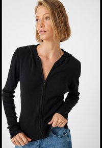 Svart ribbad zip-up hoodie med en figursydd design. Har en huva och långa ärmar. Materialet verkar mjukt och texturerat. Matchad med blå jeans.