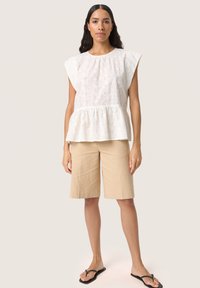 Vit blommönstrad peplumtopp med kort ärm, parat med beige knälånga shorts och svarta flip-flops. Neutral bakgrund.