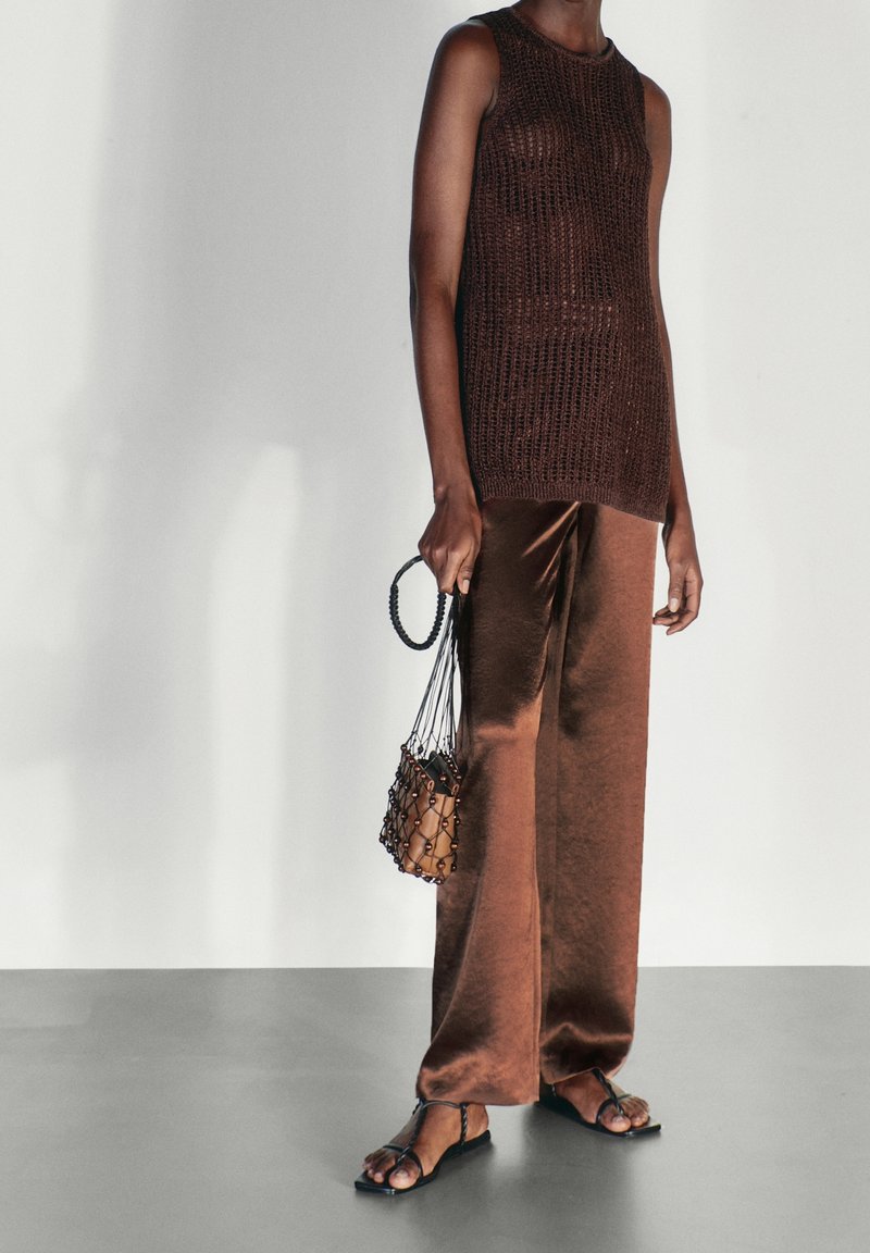 Haut en maille marron sans manches associé à un pantalon large marron brillant, des sandales noires à brides, et un petit sac en filet avec une pochette intérieure marron.
