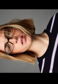 Fekete, kerek keretes szemüveg, világosbarna haj, és tengerészkék póló rózsaszín és fehér vízszintes csíkokkal. A bőrön látható szeplők vannak.