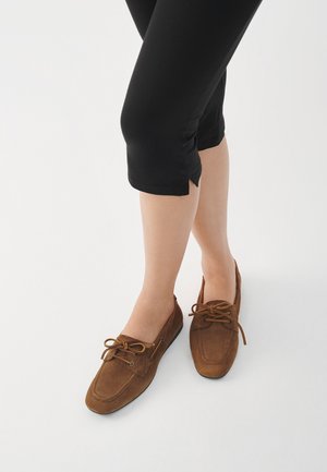 Rjavi nubuk mokasini z zaobljenim nosom, zavezovanjem in nizkim profilom. Noseni z črnimi skrajšanimi legicami na belem ozadju.