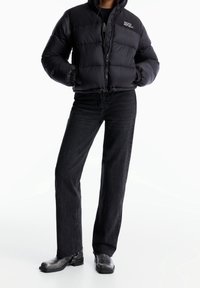 Veste matelassée noire avec fermeture éclair à l'avant et logo brod é, associée à un jean noir taille haute. Des bottines noires élégantes complètent le look.