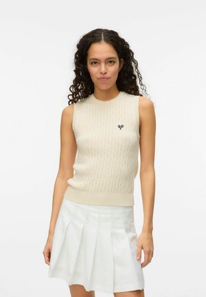 Beige mouwloos gebreid vest met ribbelstructuur en een ronde hals. Bevat een klein zwart embleem op de linkerborst. Gecombineerd met een witte geplooide rok.