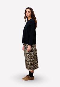 Femme aux longs cheveux ondulés portant un haut à manches longues noir, une jupe midi à imprimé léopard, des chaussettes noires et des chaussures marron, se tenant de profil.